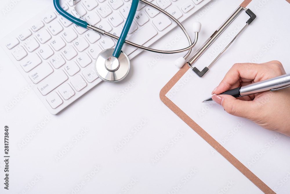 Female doctor writing a medical record case over clipboard on white working table with stethoscope, computer keyboard. Top view, flat lay, copy space Female doctor writing a medical record case over clipboard on white working table with stethoscope, computer keyboard. Top view, flat lay, copy space