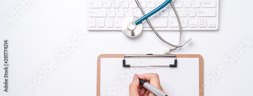 Female doctor writing a medical record case over clipboard on white working table with stethoscope, computer keyboard. Top view, flat lay, copy space photo