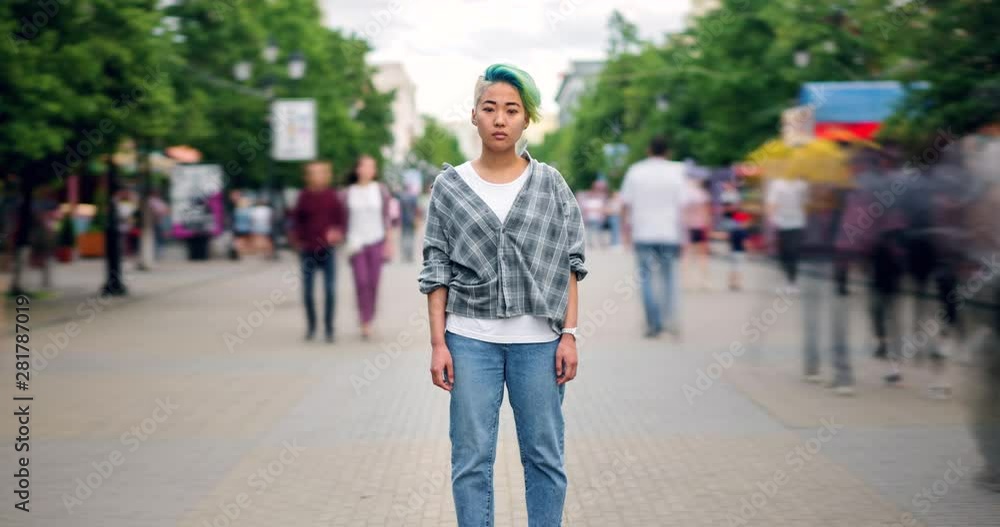 Zoom out time lapse portrait of stylish Asian teen standing in pedestrian street alone and looking at camera with serious face while people are moving around. Zoom out time lapse portrait of stylish Asian teen standing in pedestrian street alone and looking at camera with serious face while people are moving around.