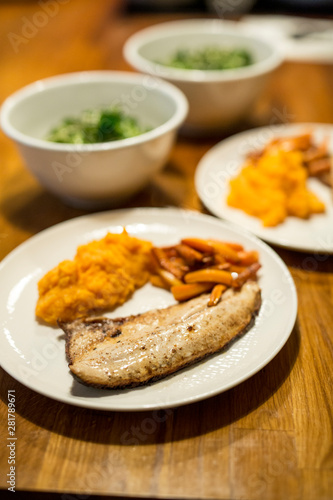 Fischfilet am Teller mit Möhren, Süsskartoffel-Stampf, Feldsalat in Schüssel, auf Holztisch, Nahaufnahme