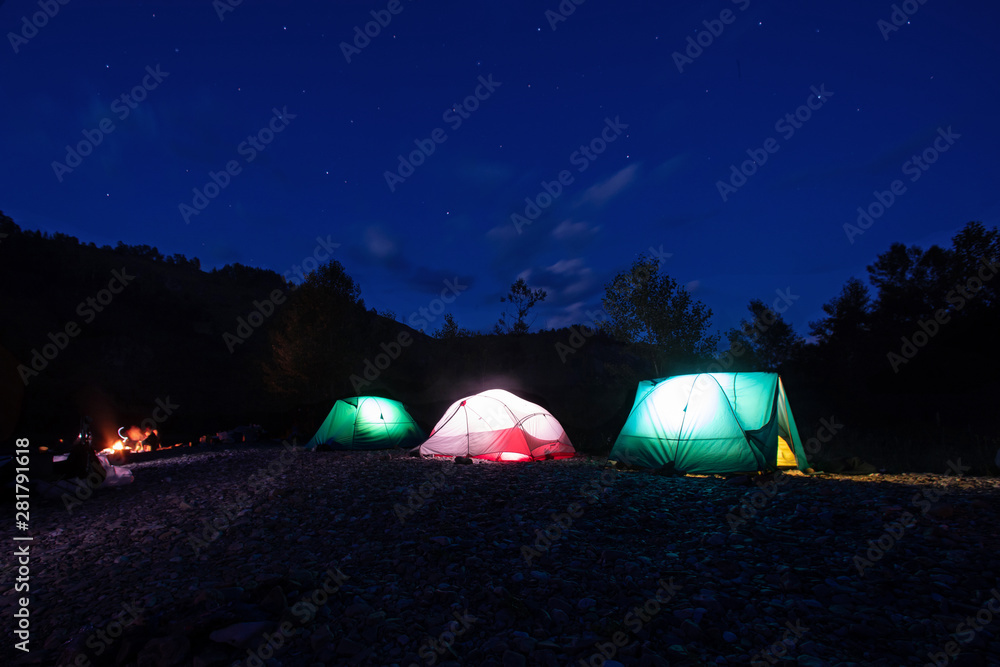 Night camping. Lighted tents and bonfire in the mountains under the night sky with stars. near the forest