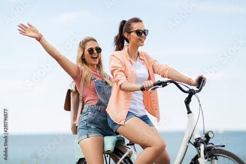 leisure and friendship concept - happy smiling teenage girls or friends riding bicycle together at seaside in summer