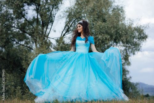 Portrait fashion girl standing near tree. Amazing brown hair model in blue dress looking at camera and posing. Fairytale about young princess.Perfect make up. Young elegant woman in blue long flying