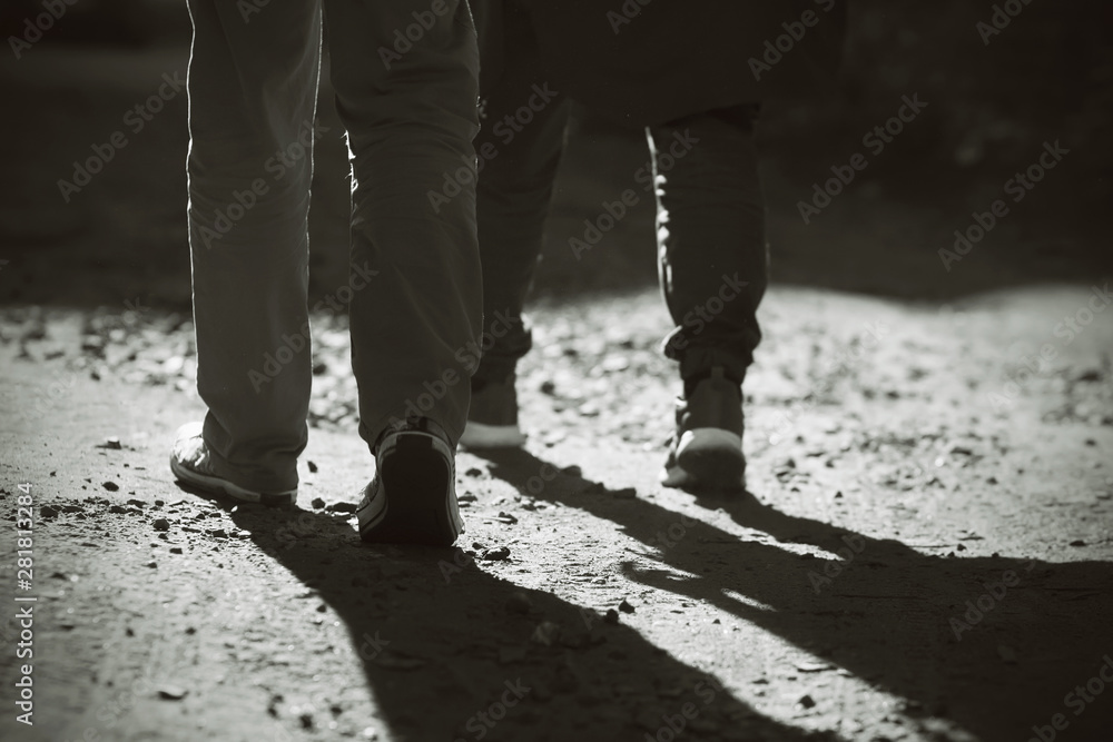 Black-and-white image of the, where its feet two man, dressed in sneakers and trousers, go on foot on sandy-rocky road. People go from light to darkness. Black-and-white image of the, where its feet two man, dressed in sneakers and trousers, go on foot on sandy-rocky road. People go from light to darkness.