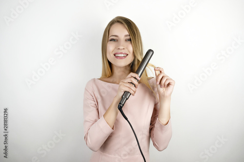 young beautiful blonde woman wearing trendy pink dress using hair streightener to make fashionable hairstyle by herself on isolated white background