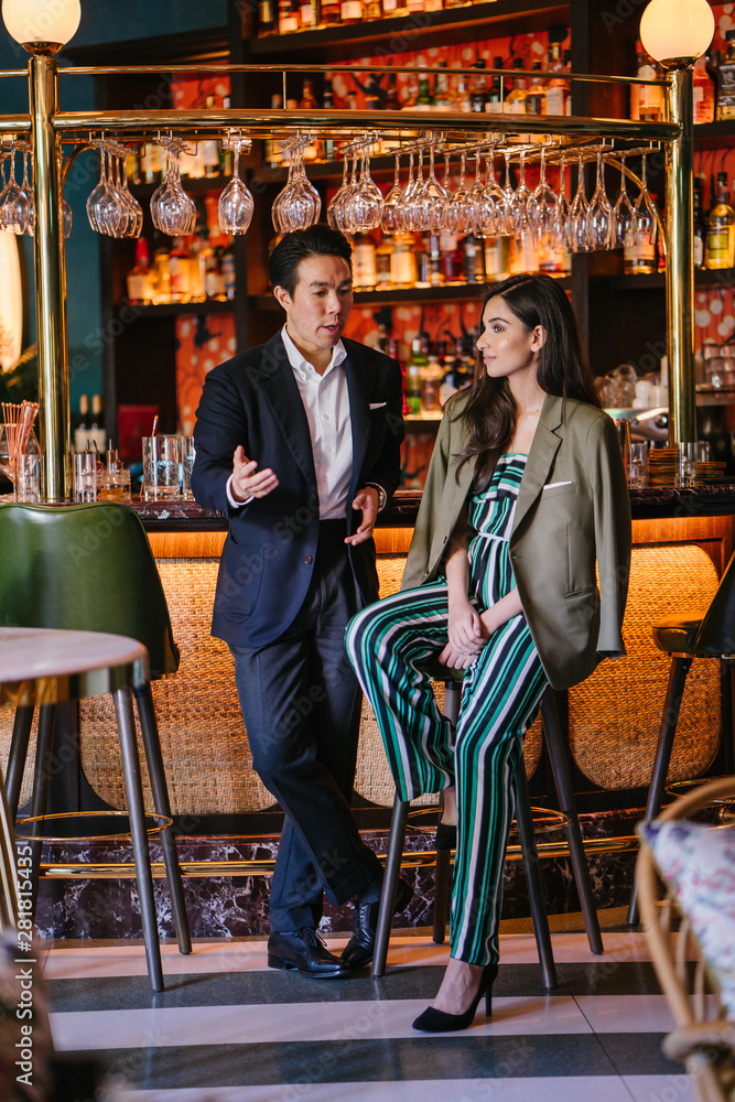 Portrait of two professionally and well-dressed young Asian individuals out on a date at a classy bar together. One is a Chinese man in a suit with his elegant Indian woman date -- they are talking.