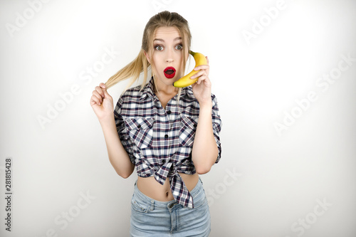 young beautiful blonde woman wearing trendy checkered shirt and denim shorts holding banana like a telephone isolated white background