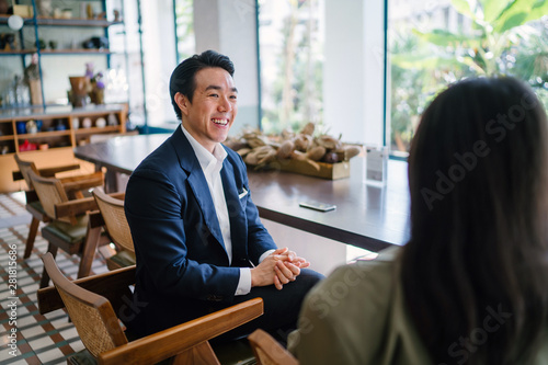 A young and attractive Chinese Asian man in a well-fitted navy suit and pocket square is talking to his companion in a meeting. He is gesturing as he talks to her in a trendy cafe or office.