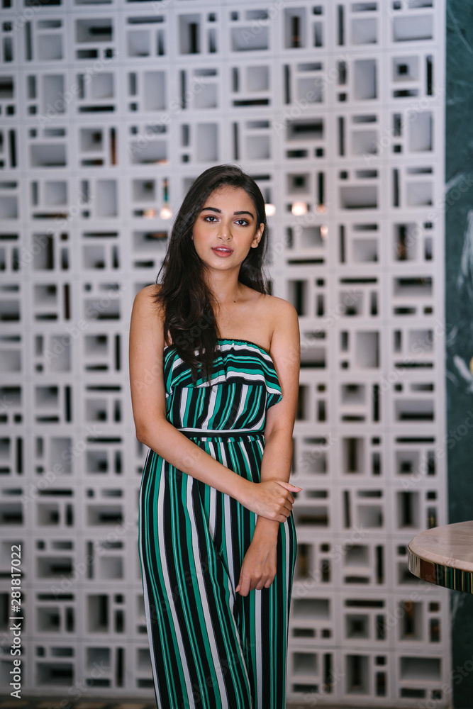 Portrait head shot of a young, attractive, beautiful and stylish Indian Asian woman in a green striped jumpsuit. She is smiling happily as she poses for her portrait in a stylish interior. Portrait head shot of a young, attractive, beautiful and stylish Indian Asian woman in a green striped jumpsuit. She is smiling happily as she poses for her portrait in a stylish interior.