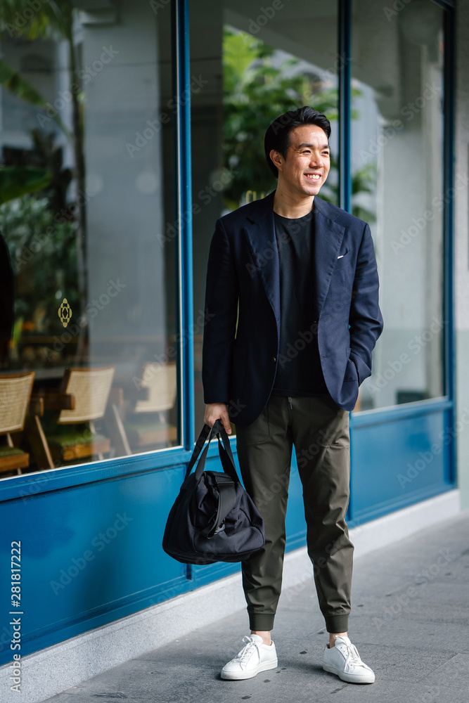 Φωτογραφία A young Chinese Asian man in a stylish casual outfit is holding his gym bag while standing by the street in Asia