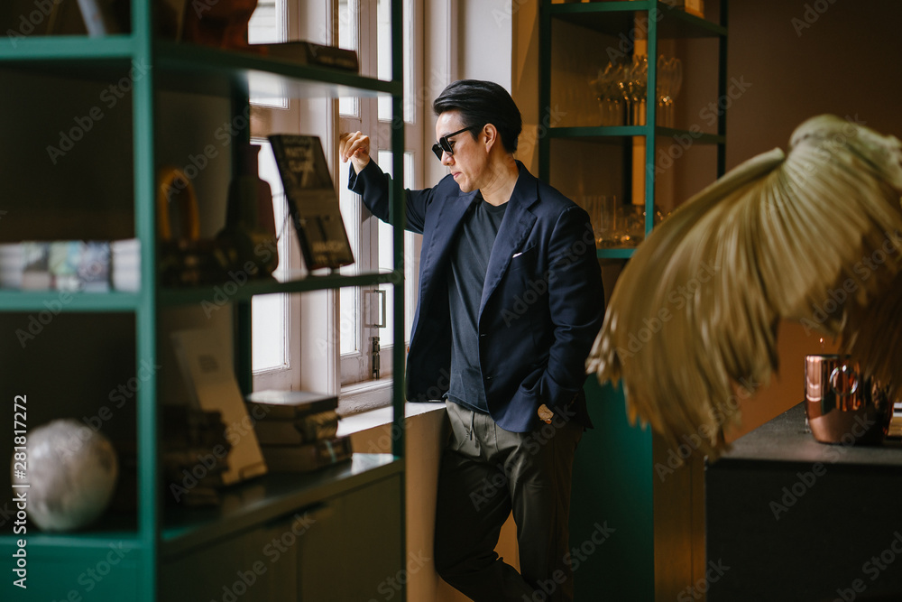 Portrait of a handsome, good looking young Chinese Asian man in a casual and well-tailored suit, tee shirt and sunglasses as he gazes out the window of a well-decorated room during the day.