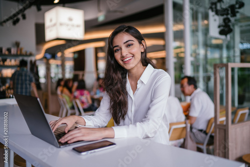 Portrait of young, beautiful and hopeful Indian Asian woman in a casual white shirt as she talks on her smartphone. She is taking a break from working on her laptop in a cafe of coworking space.