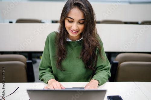 Photography Portrait of a beautiful, young and intelligent-looking Indian Asian woman student wearing a white shirt and green tracker smiling as she works on her laptop in a university classroom