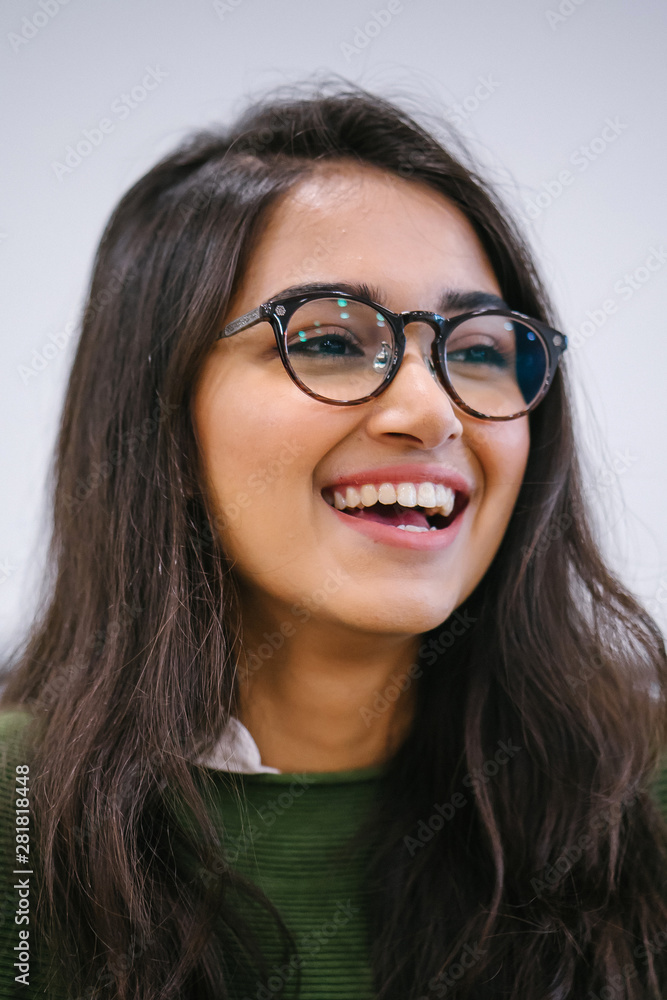 A close up head shot portrait of a preppy, young, beautiful, confident and attractive Indian Asian woman in a green sweater and spectacles against a white background. She is smiling happily. A close up head shot portrait of a preppy, young, beautiful, confident and attractive Indian Asian woman in a green sweater and spectacles against a white background. She is smiling happily.