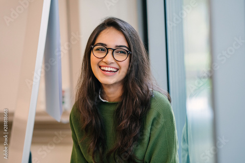 Photo A close up head shot portrait of a preppy, young, beautiful, confident and attractive Indian Asian woman in a green sweater and spectacles in a classroom or office