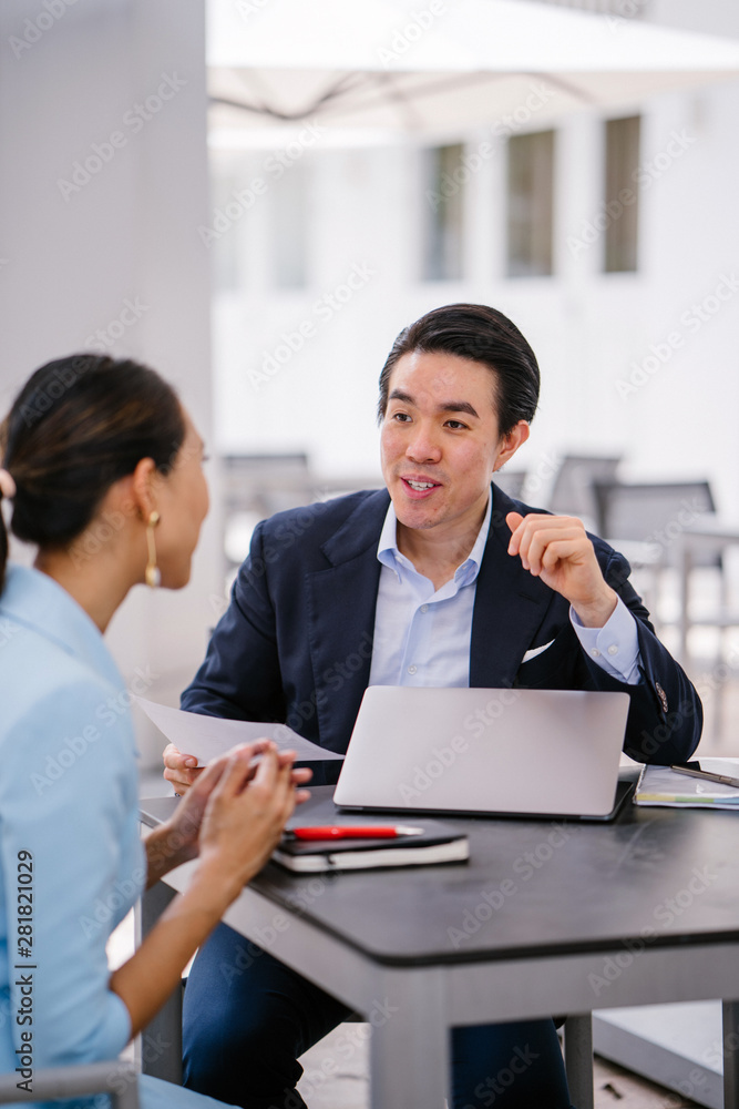 A Chinese Asian manager in a suit has a meeting with his colleague, a woman in a pale blue suit. He is conducting a performance appraisal during this meeting. They are smiling and talking.