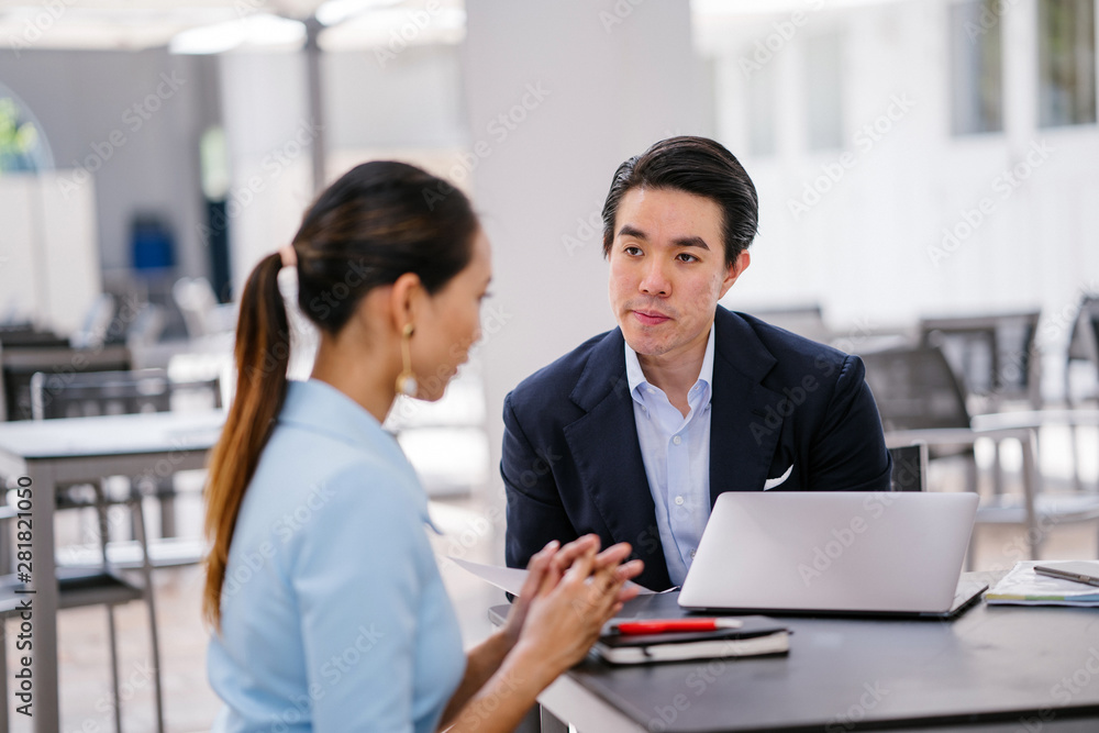 A Chinese Asian manager in a suit has a meeting with his colleague, a woman in a pale blue suit. He is conducting a performance appraisal during this meeting. They are smiling and talking. A Chinese Asian manager in a suit has a meeting with his colleague, a woman in a pale blue suit. He is conducting a performance appraisal during this meeting. They are smiling and talking.