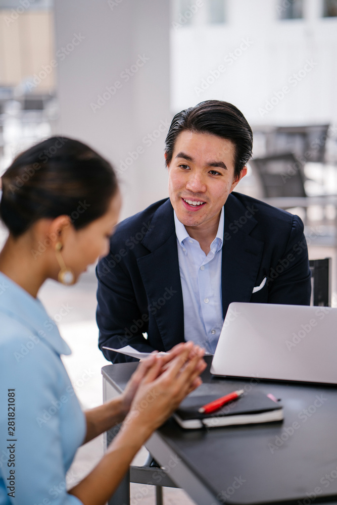 Fototapeta premium A Chinese Asian manager in a suit has a meeting with his colleague, a woman in a pale blue suit. He is conducting a performance appraisal during this meeting. They are smiling and talking.