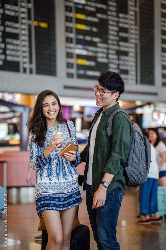 Portrait of a young and casually dressed Asian interracial couple (Korean man and his Indian girlfriend) at the airport. They are smiling and laughing together, excited about their vacation.