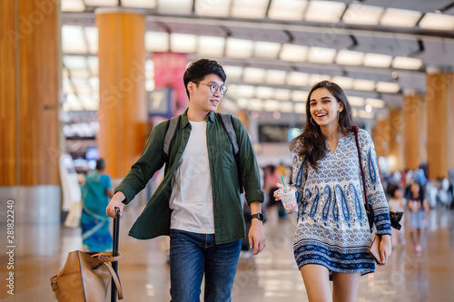 A young and photogenic Asian couple (Korean man, Indian girlfriend) smile as they walk in a futuristic airport. They are pulling their luggage behind them as they look for their check in counter.