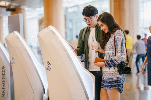 A portrait of a pair of young and excited Asian travelers (Korean man and his Indian friend) checking in to board their plane for their holiday at an automated check-in booth in the airport. © Danon