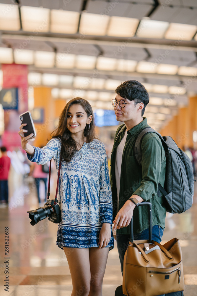 A young and diverse Asian couple (Korean man and his Indian girlfriend) take a selfie together in a new country. They have just arrived in a new country for a tour and are excited.