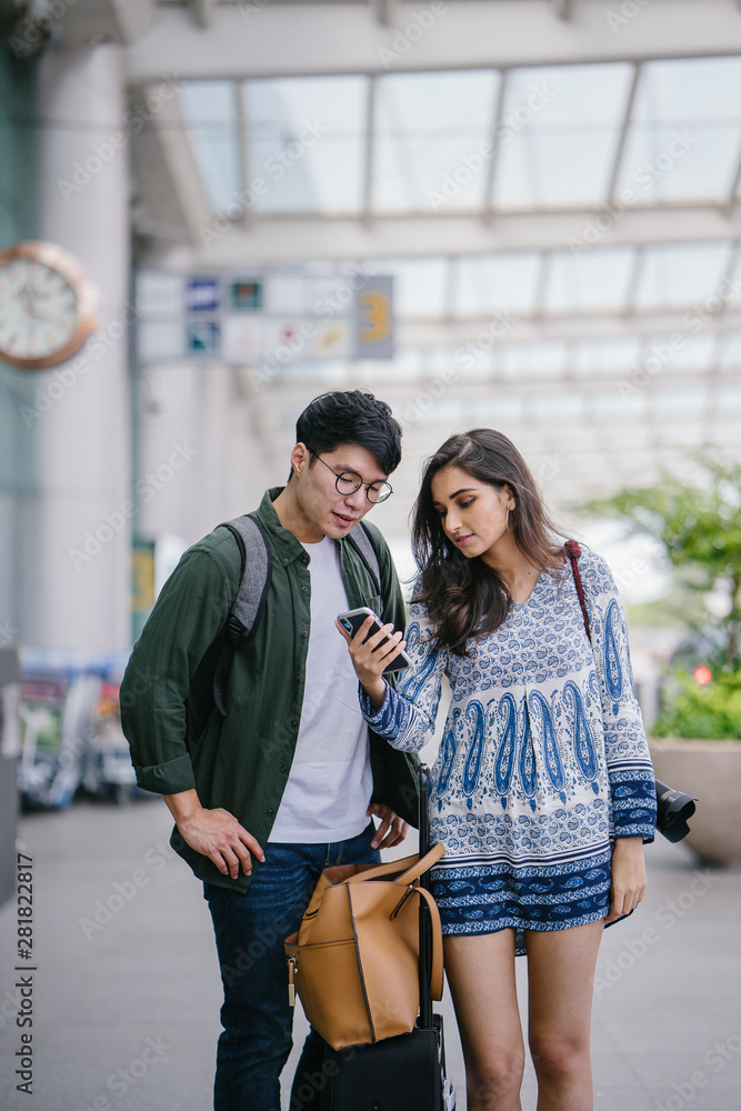 A young and diverse Asian couple (Korean man and his Indian girlfriend) is booking a ride on their ride-hailing app on her smartphone. They are waiting at the cab stand in the airport.