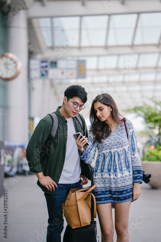 A young and diverse Asian couple (Korean man and his Indian girlfriend) is booking a ride on their ride-hailing app on her smartphone. They are waiting at the cab stand in the airport.