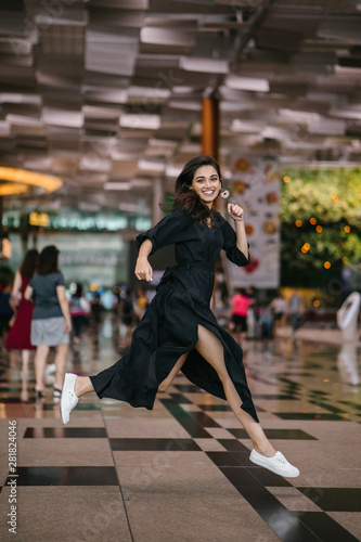 Portrait of an attractive  young and athletic north Indian Asian woman smiling and laughing as she leaps and jumps indoors in a mall or airport. She is wearing a casual black flowing dress.
