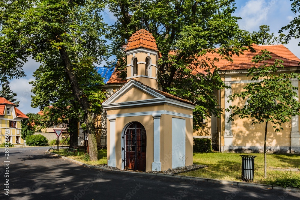 Panensky Tynec, Czech Republic - July 15 2019: Chapel of John of Nepomuk with yellow and white facade standing in the street. Green grass, trees around. Sunny summer day with blue sky and clouds.