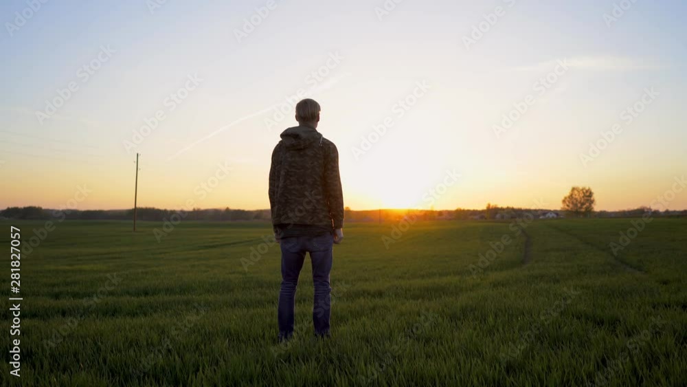 View from the back of a young man standing in the middle of a green field watching towards the horizon in a beautiful sunset View from the back of a young man standing in the middle of a green field watching towards the horizon in a beautiful sunset