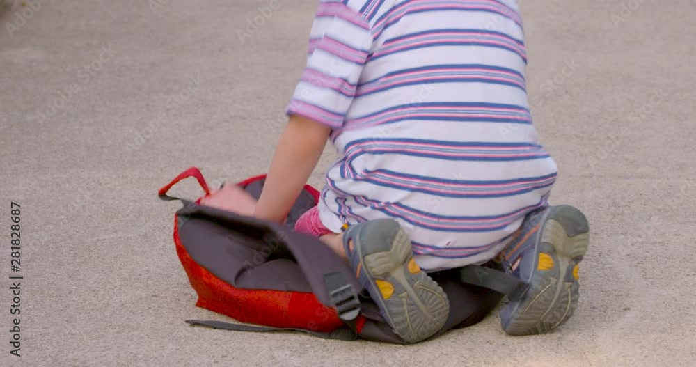 A little boy falls on to his backpack, then picks it up and puts it on his back A little boy falls on to his backpack, then picks it up and puts it on his back