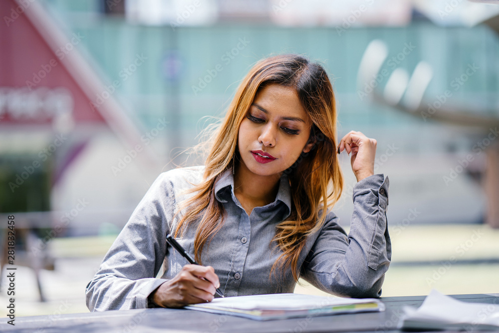 Portrait of a tanned and professional young Southeast Asian business woman in a collared shirt sitting on a bench in the city. She is smiling as she writes and jots in her notebook. Portrait of a tanned and professional young Southeast Asian business woman in a collared shirt sitting on a bench in the city. She is smiling as she writes and jots in her notebook.