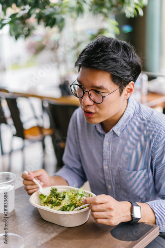 A Korean Asian man enjoys his salad lunch as he sits in a cafe in the city during the day. He is tall  handsome and is wearing a collared shirt and spectacles.