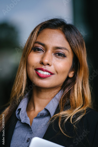 A close-up portrait of a young, tanned, beautiful and confident Southeast Asian woman in a business suit. She has a radiant smile and dyed hair and is looking intently at the camera.