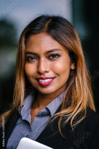 A close-up portrait of a young, tanned, beautiful and confident Southeast Asian woman in a business suit. She has a radiant smile and dyed hair and is looking intently at the camera.