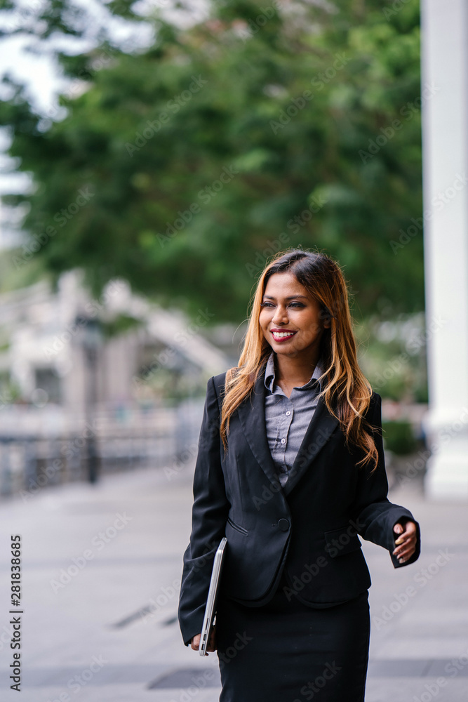 Portrait of a beautiful, young and attractive Southeast Asian woman in a business suit walking in a city in Asia during the day with her laptop computer. She is smiling confidently.