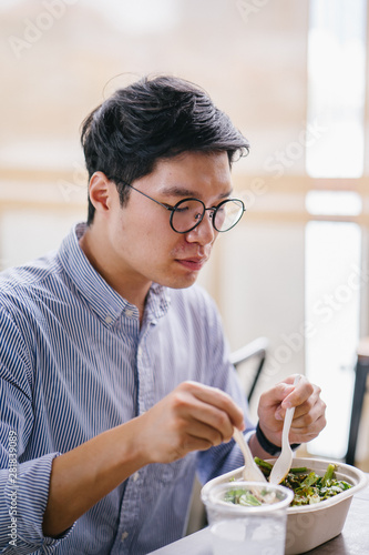 A Korean Asian man enjoys his salad lunch as he sits in a cafe in the city during the day. He is tall, handsome and is wearing a collared shirt and spectacles.