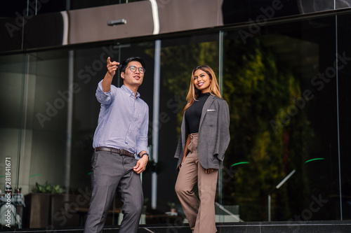 A tall, confident and clean-cut Asian man in shirt and pants smiles as he gives a tour or directions to the city to a young and beautiful Malay Asian woman in a suit during the day.