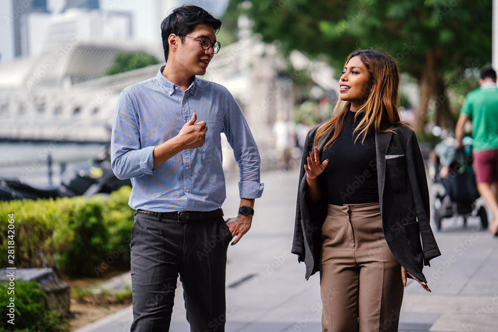 Portrait of two diverse Asian business people (colleagues meeting for lunch) walking in the city (Singapore River). One is a Korean man, the other a Malay woman. They are both smiling as they chat. Portrait of two diverse Asian business people (colleagues meeting for lunch) walking in the city (Singapore River). One is a Korean man, the other a Malay woman. They are both smiling as they chat.