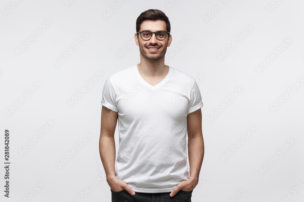 Handsome smiling man in white t-shirt and trendy eyeglasses, standing with hands in pockets, isolated on gray background Handsome smiling man in white t-shirt and trendy eyeglasses, standing with hands in pockets, isolated on gray background