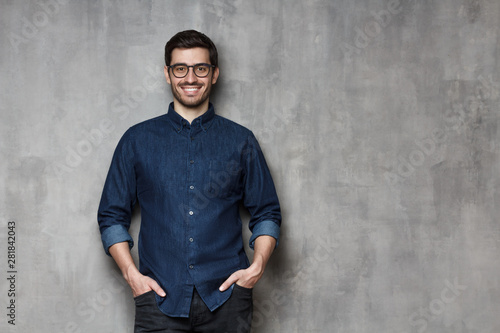 Young handsome man wearing trendy glasses and denim shirt standing leaning on gray textured wall with copy space on right side