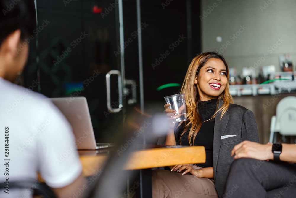 Portrait of a beautiful, fashionable, millennial Asian Malay businesswoman smiling to her colleague whose out of view and is sitting in a cafeteria while holding her cup of beverage during the day. Portrait of a beautiful, fashionable, millennial Asian Malay businesswoman smiling to her colleague whose out of view and is sitting in a cafeteria while holding her cup of beverage during the day.