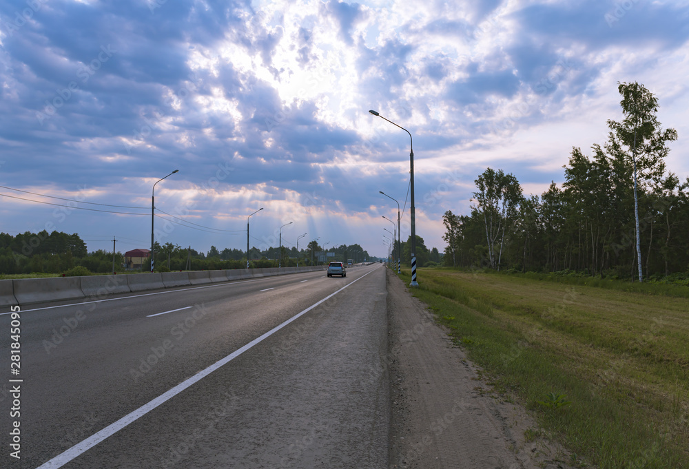 The car rides on a suburban highway surrounded by trees. The sun's rays make their way through the beautiful blue clouds in the sky. Landscape