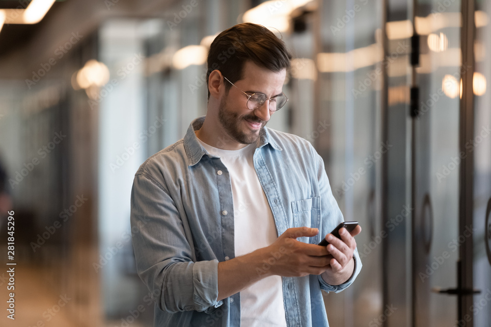 Happy young businessman using smart phone standing in office hallway Happy young businessman using smart phone standing in office hallway