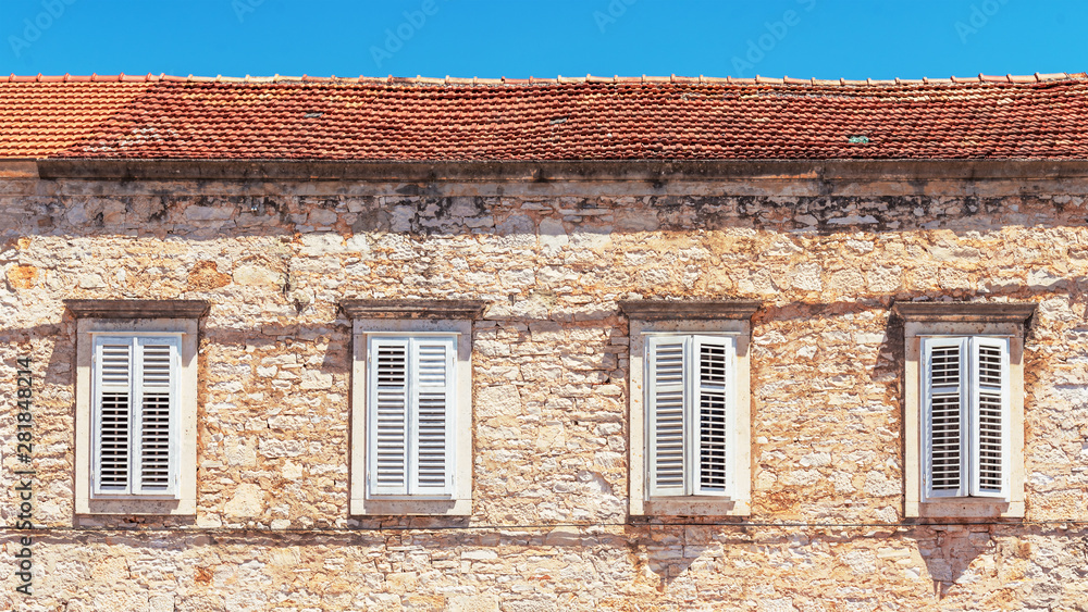 Fragment of an old stone building with four windows, white shutters, blue sky as background