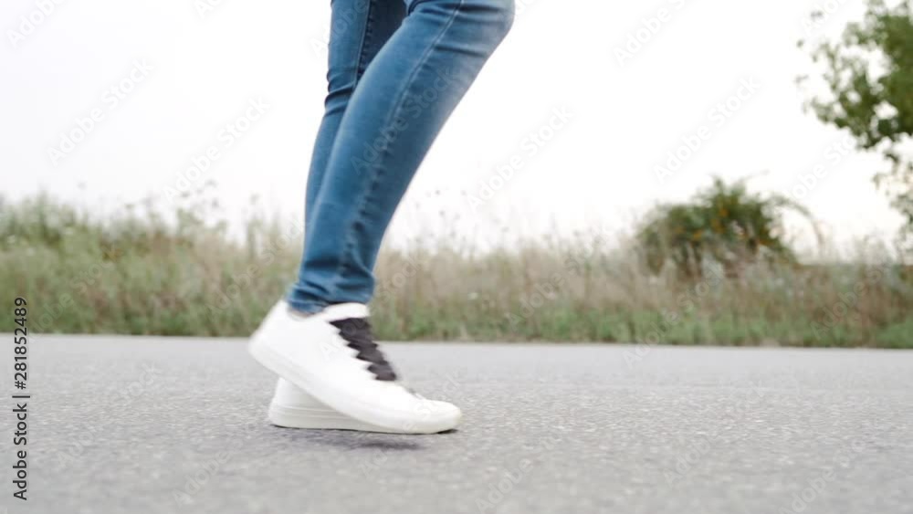 Unrecognizable woman in jeans and white sneakers walks along on asphalt road ahead. Concept of modern shoes, travel, hiking in summer nature.Camera shoots from right side. Unrecognizable woman in jeans and white sneakers walks along on asphalt road ahead. Concept of modern shoes, travel, hiking in summer nature.Camera shoots from right side.