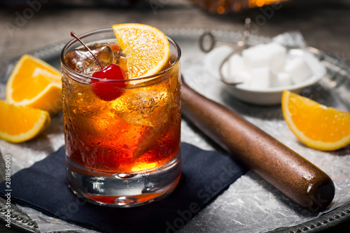 Old Fashioned Cocktail On Ice with Cherry and Orange Garnish, Sugar Cubes, and Muddler on Tray