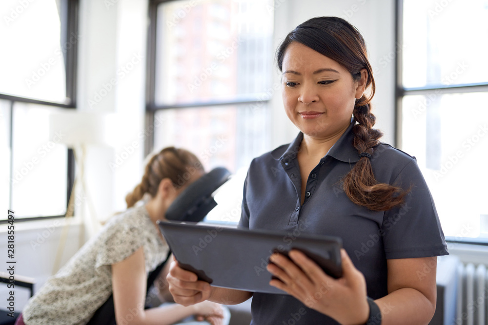 portrait of a Chinese woman massage therapist holding a tablet while filling the file of an attractive blond client at her workplace in a bright office portrait of a Chinese woman massage therapist holding a tablet while filling the file of an attractive blond client at her workplace in a bright office