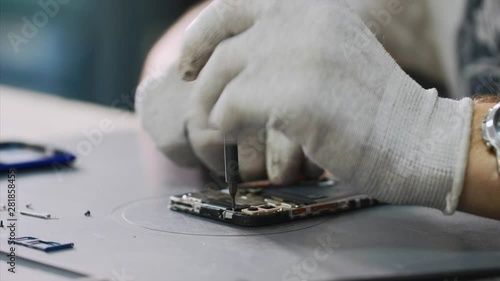 Repairing mobile phone in workshop, hands closeup. Repairman in white gloves disassemble smartphone with screwdriver in service center. Master in white gloves unscrews bolts. Repair and maintenance.